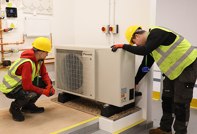 Students wearing hard hats and hi-vis jackets next to Air Source Heat Pump