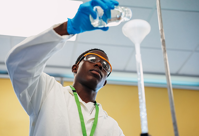 Student pouring liquid into a funnel whilst wearing gloves, safety goggles and an apron 